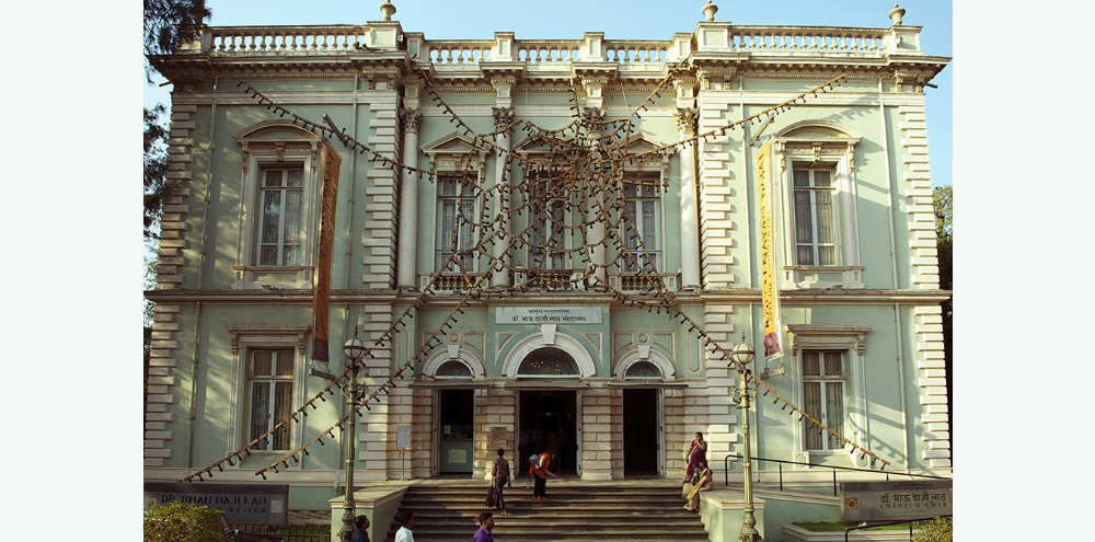 An installation of rubber stamps strung together and arranged in a cobweb-like manner on the facade of a blue building.