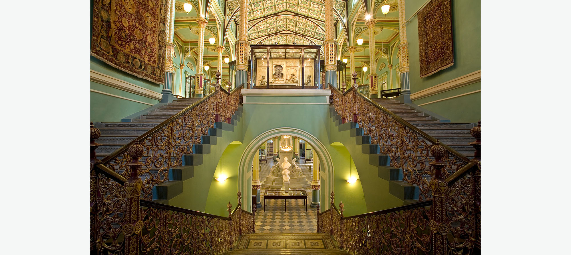 A brightly lit staircase with blue walls, ornate railings and textiles displayed on the walls.