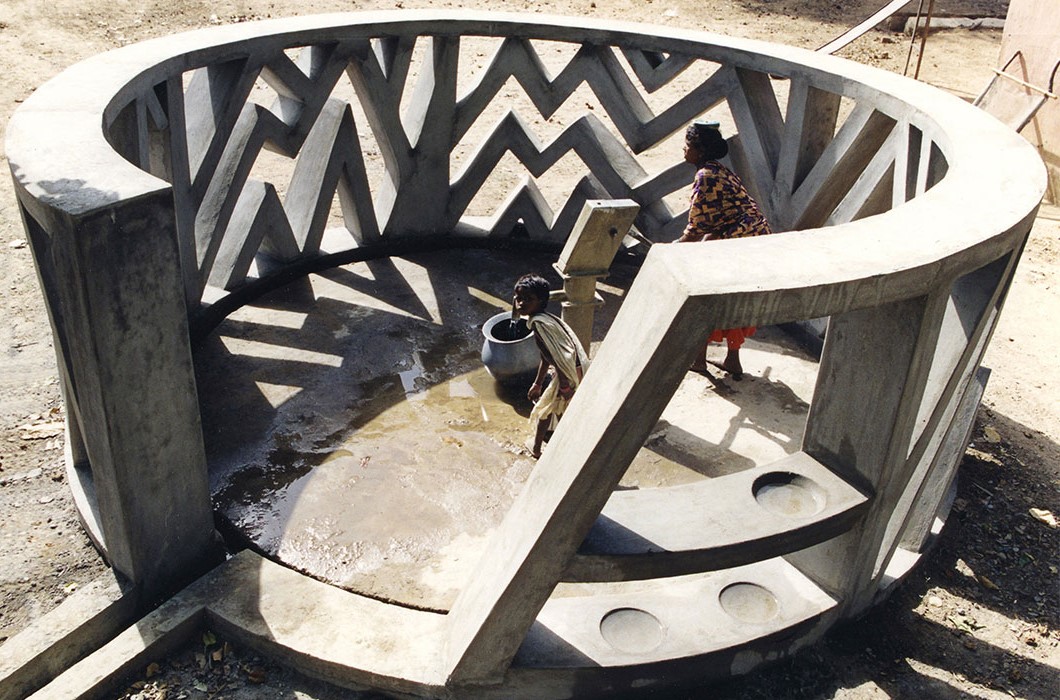 A circular wall with irregular chevron-striped openings surrounds a hand-operated water pump where a woman and child collect water.